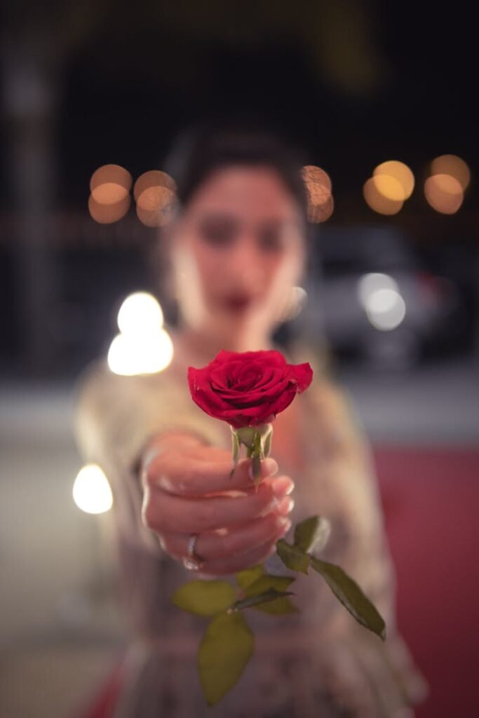 Blurry woman holding a vibrant red rose in focus against a bokeh-lit background.